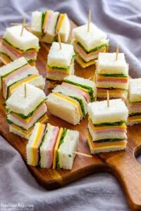 Mini tea party sandwiches with meat, cheese, lettuce, and a toothpick on a cutting board.