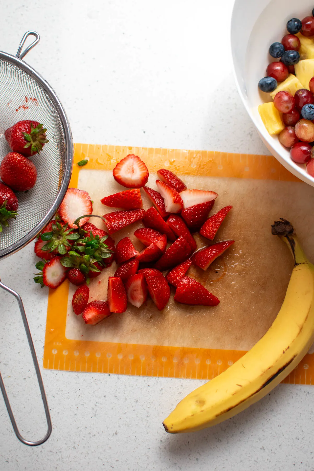 Chopped strawberries on cutting board with banana and large bowl of fruit nearby.