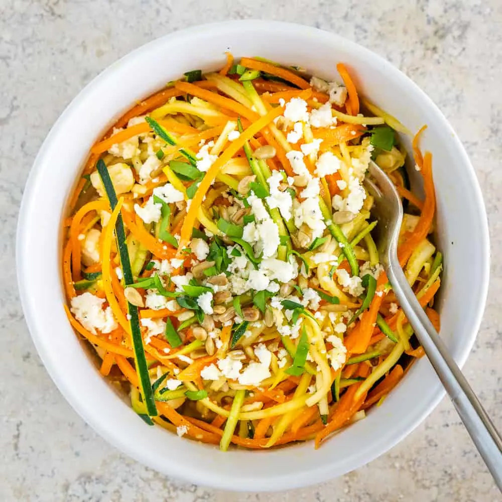 Spoon rests in spiralized carrot zucchini salad with feta in white bowl. 