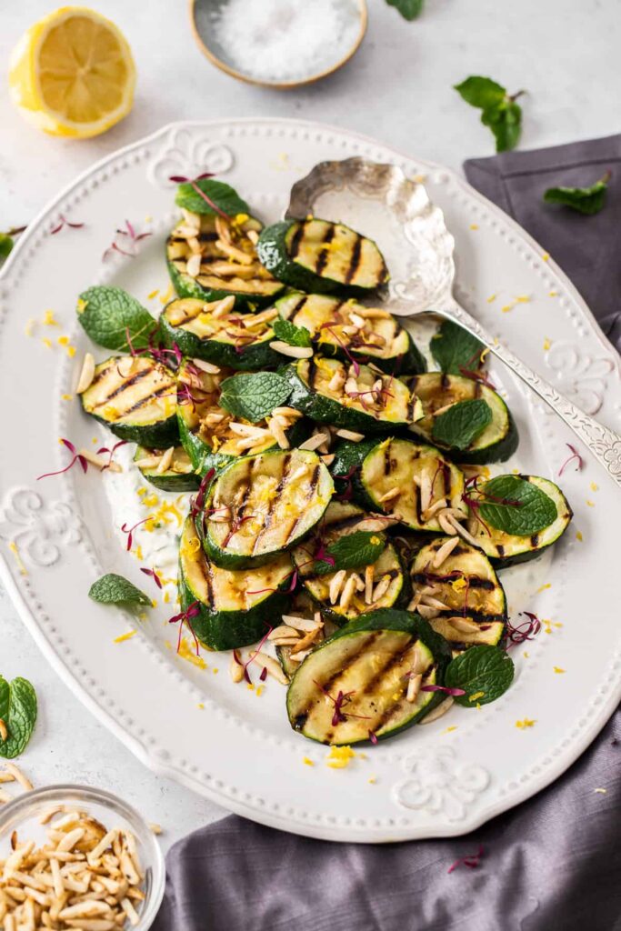 Grilled zucchini slices on a large white serving plater with a silver serving spoon.