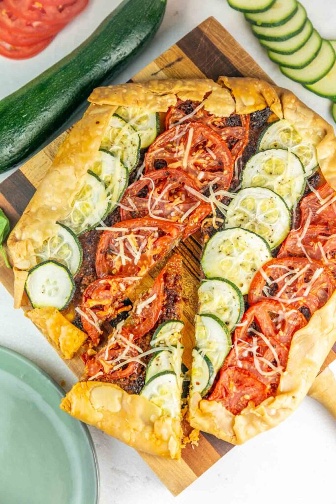 Tomato and zucchini Galette on a wooden cutting board next to fresh tomatoes and zucchini.