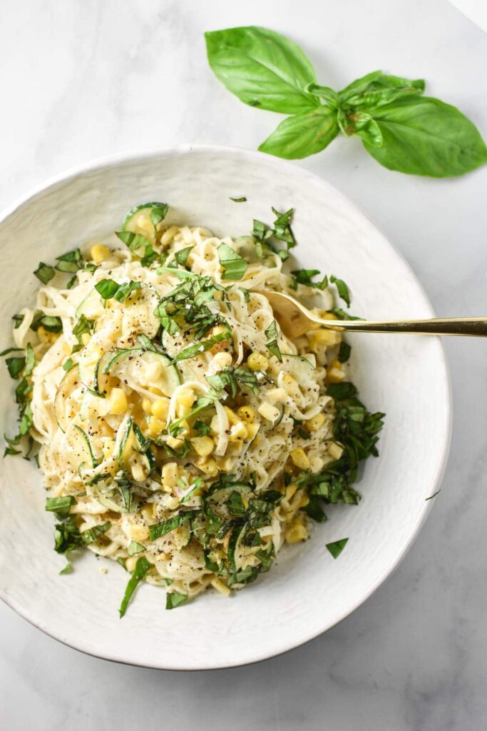 Creamy sweet corn pasta with zucchini and green garnish on a white plate.