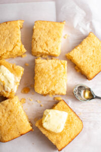 Pieces of cornbread, some with pats of butter and drizzled with honey, on piece of parchment paper.