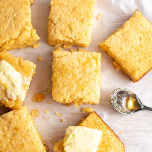 Pieces of cornbread, some with pats of butter and drizzled with honey, on piece of parchment paper.