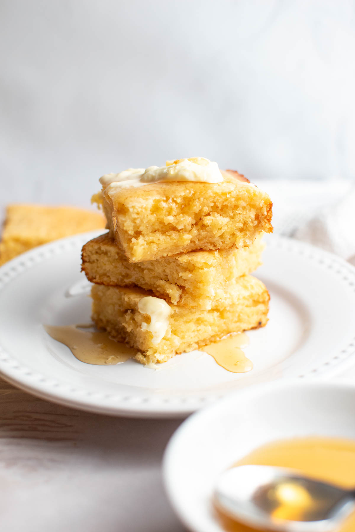 Stack of 3 pieces of cornbread with butter on top and covered in honey on white plate.
