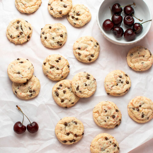 Several cherry chip cake mix cookies scattered on parchment paper with bowl of fresh cherries nearby.