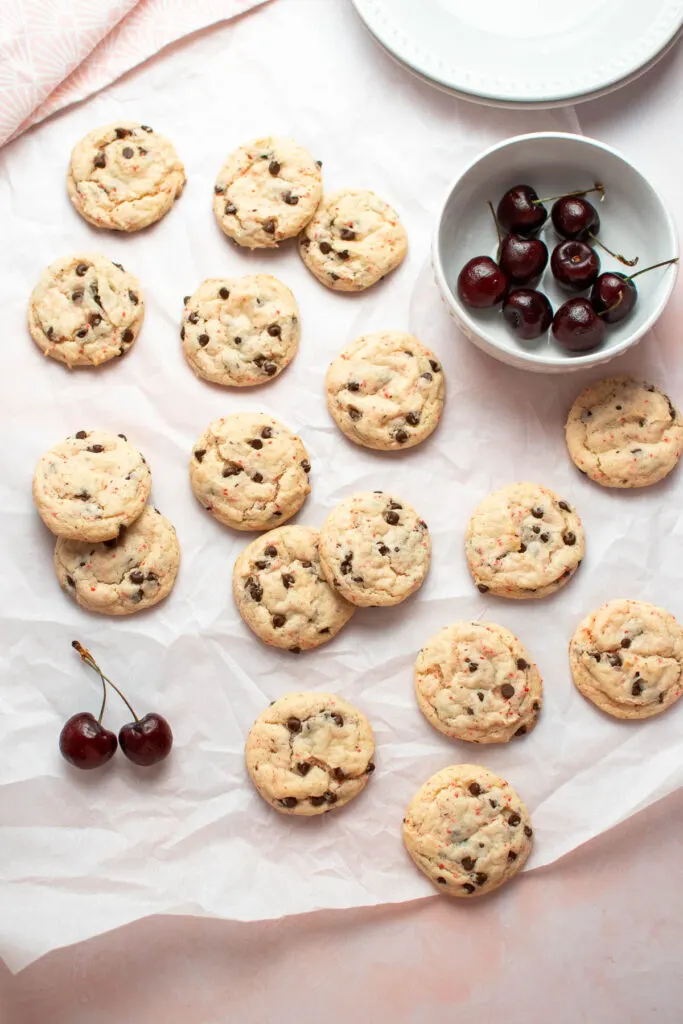Several cherry chip cake mix cookies scattered on parchment paper with bowl of fresh cherries nearby.