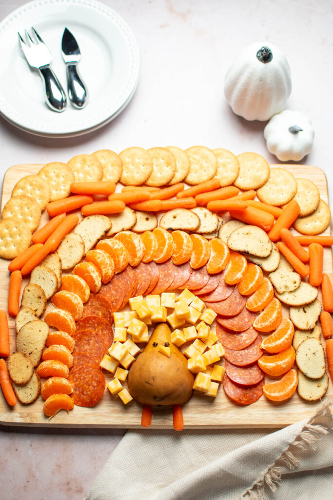 Turkey shaped charcuterie board foods on large wood cutting board with plates and pumpkins nearby.