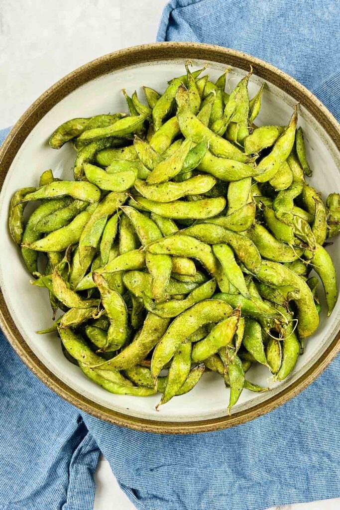 A large bowl filled with crispy roasted edamame beans on a blue kitchen towel.