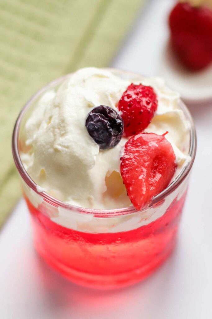 A clear glass with red jello topped with white cream and berries.