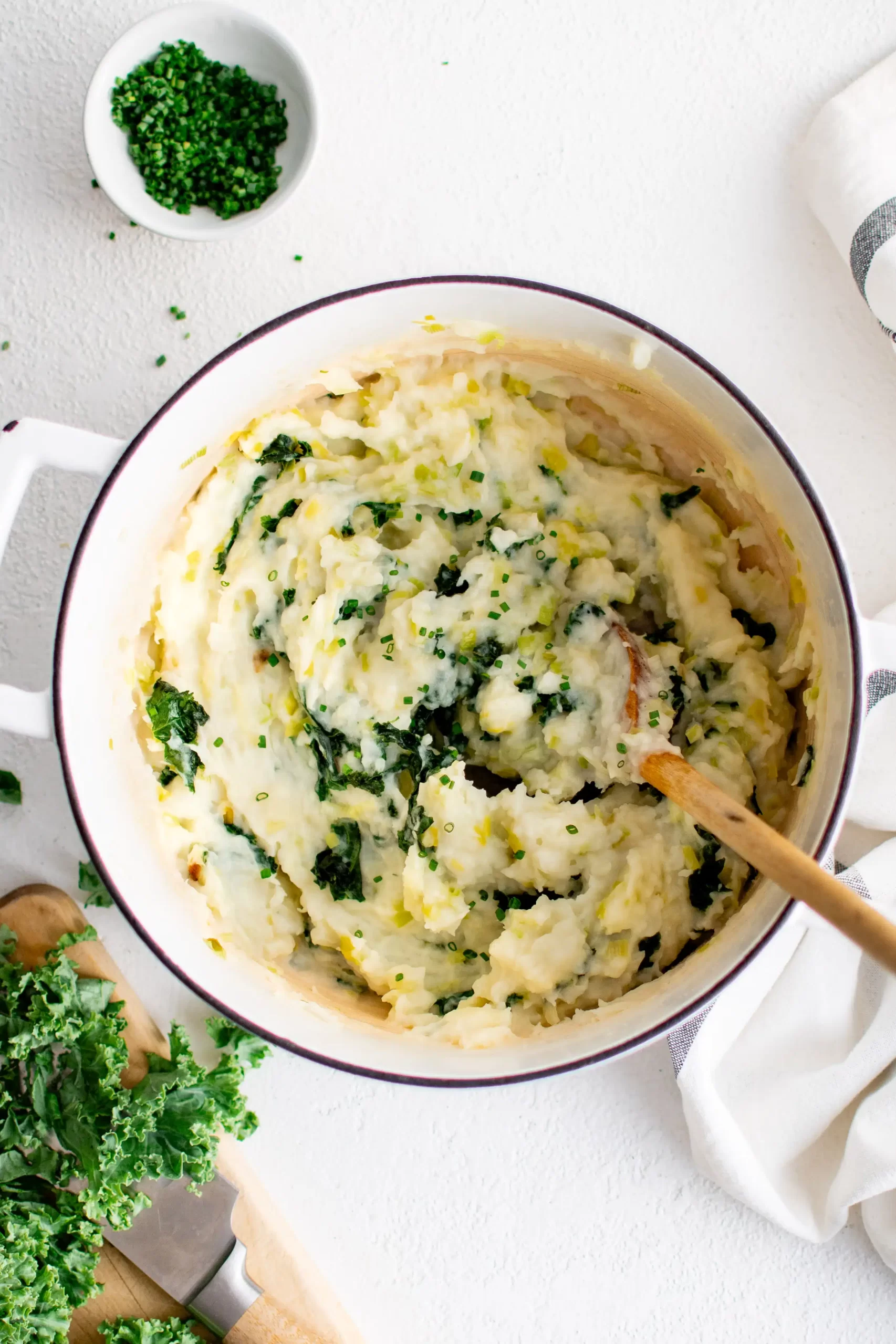 Mashed potatoes with greens mixed in in a large white heavy bottom pot with a wooden spoon.