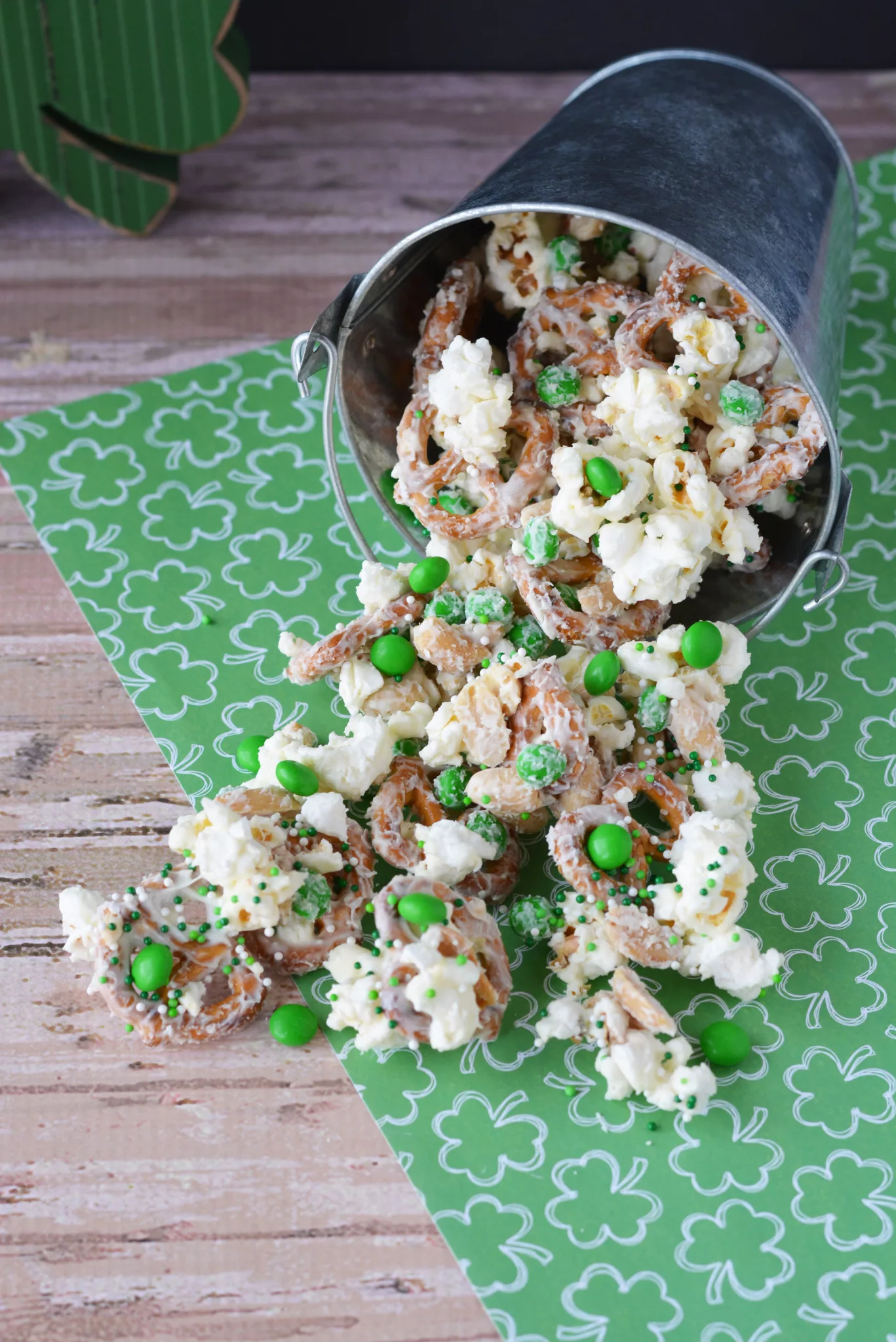 St. Patrick's Day snack mix spills from a silver bucket onto a table with a shamrock-patterned tablecloth.