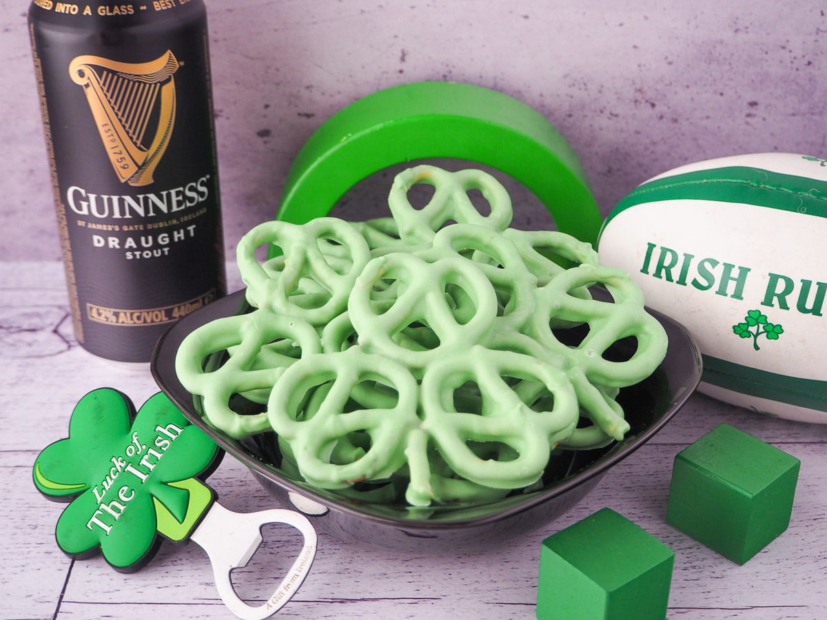 Pretzels dipped in green chocolate in the shape of shamrocks surrounded by Irish party decor.