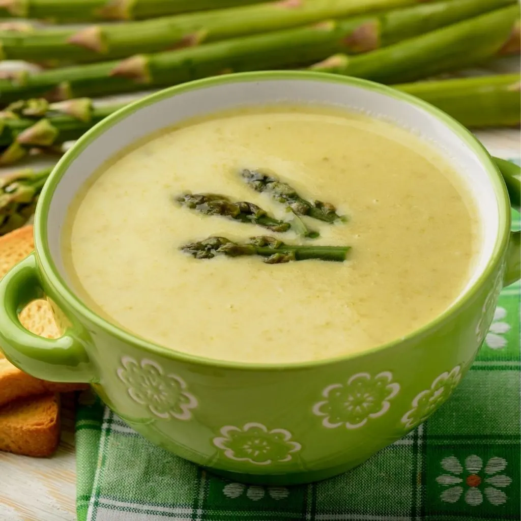 Asparagus soup with asparagus garnish in a green bowl next to fresh asparagus stalks.