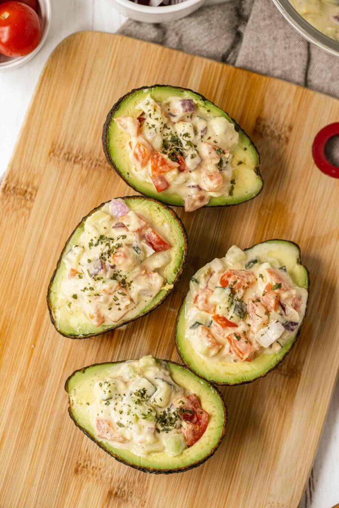 Four stuffed avocados with a creamy vegetable mixture on a wooden cutting board.