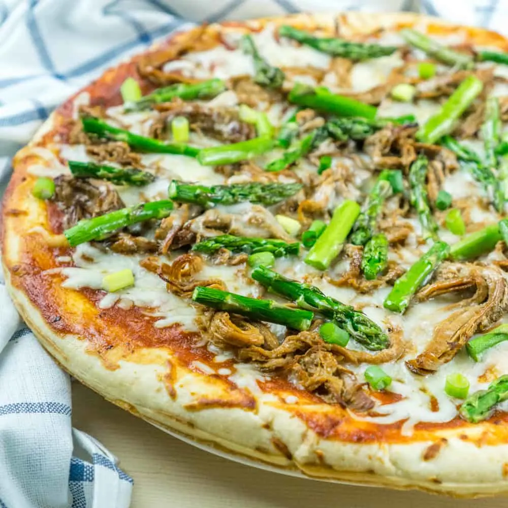 A pulled pork pizza with asparagus on top next to a white and blue kitchen towel.