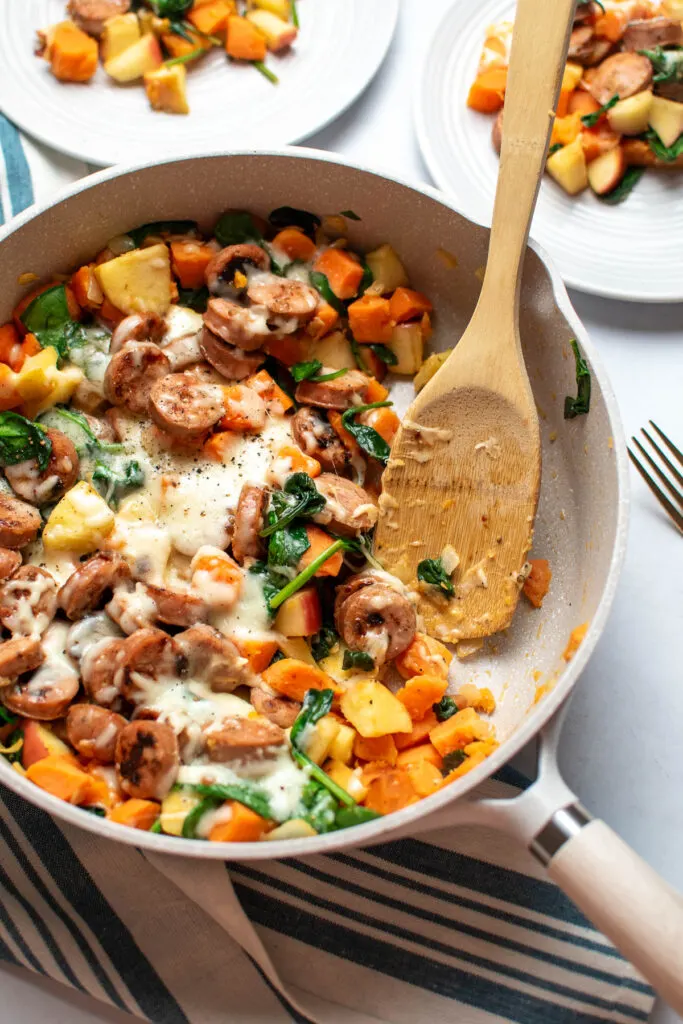 Wooden spoon rests in frying pan with chicken sausage, spinach, and sweet potato skillet.