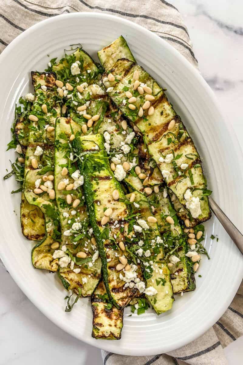 Grilled zucchini salad on a large white platter with a serving spoon.