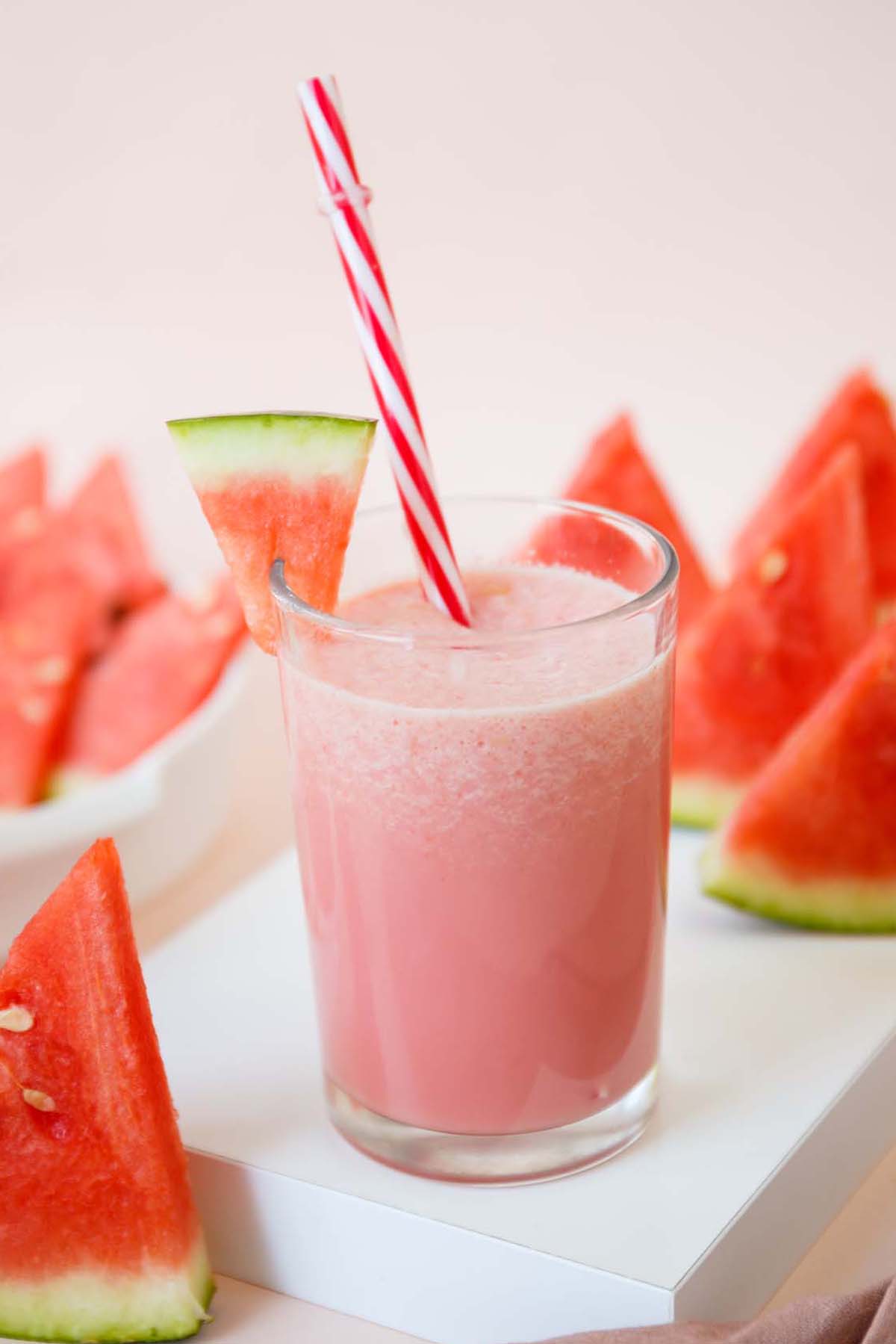 A watermelon milkshake in a glass with watermelon garnish and a red and white striped straw.