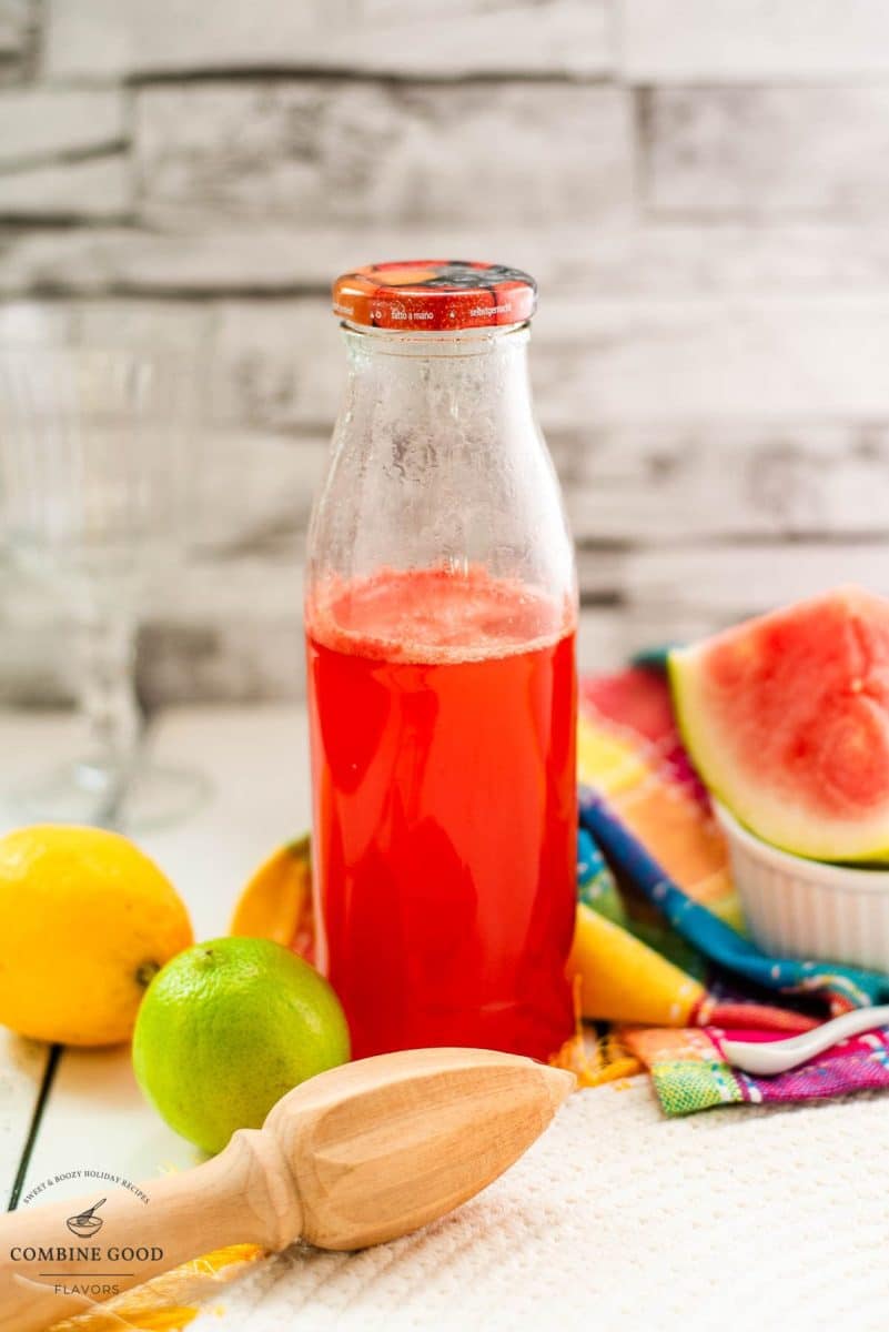 A tall thin jar of watermelon syrup next to a lemon, lime, and watermelon wedge.