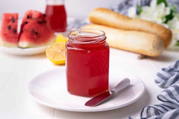 Watermelon jam in a mason gar on a white plate with a knife.