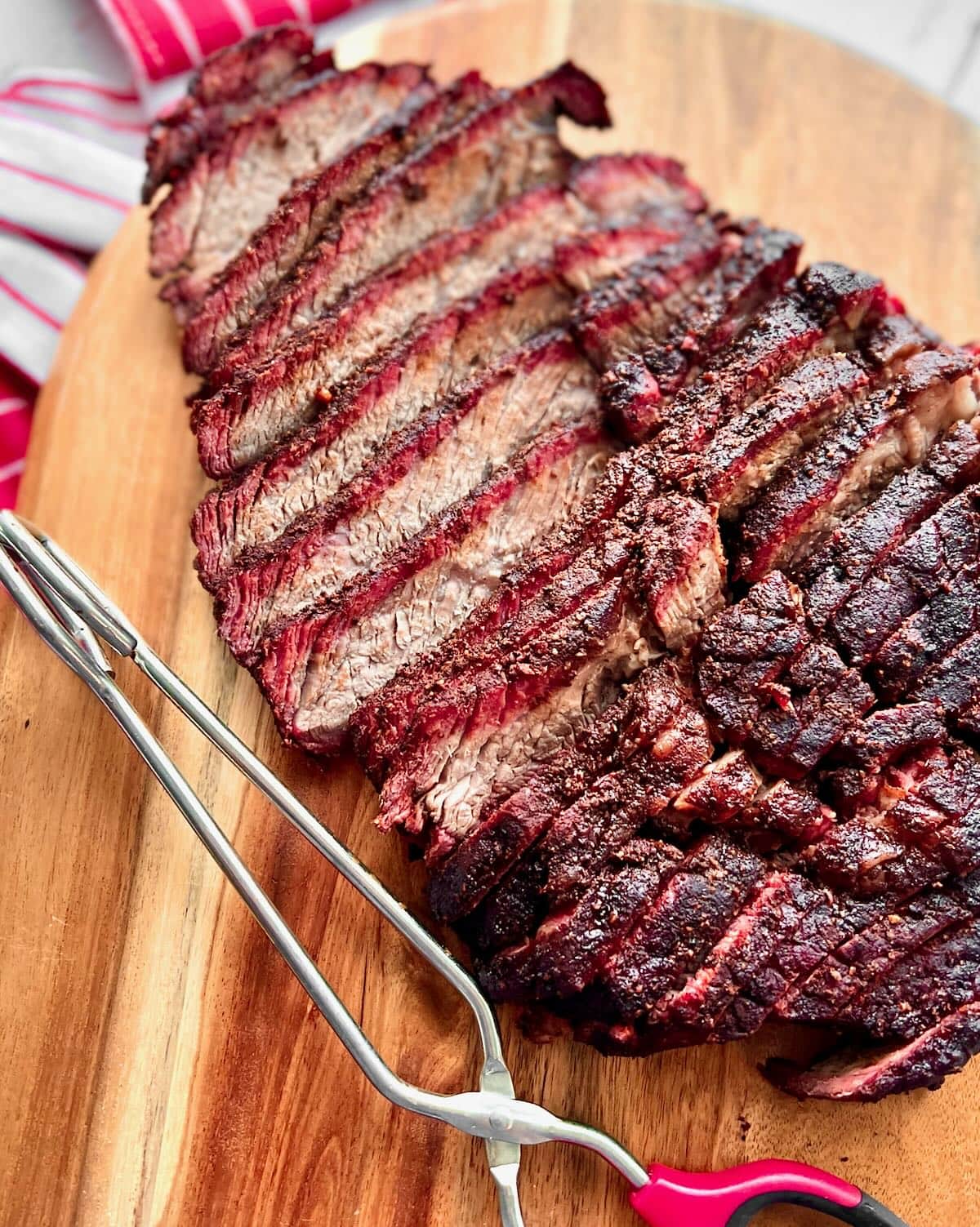Thinly sliced smoked brisket on a wooden cutting board next to tongs.
