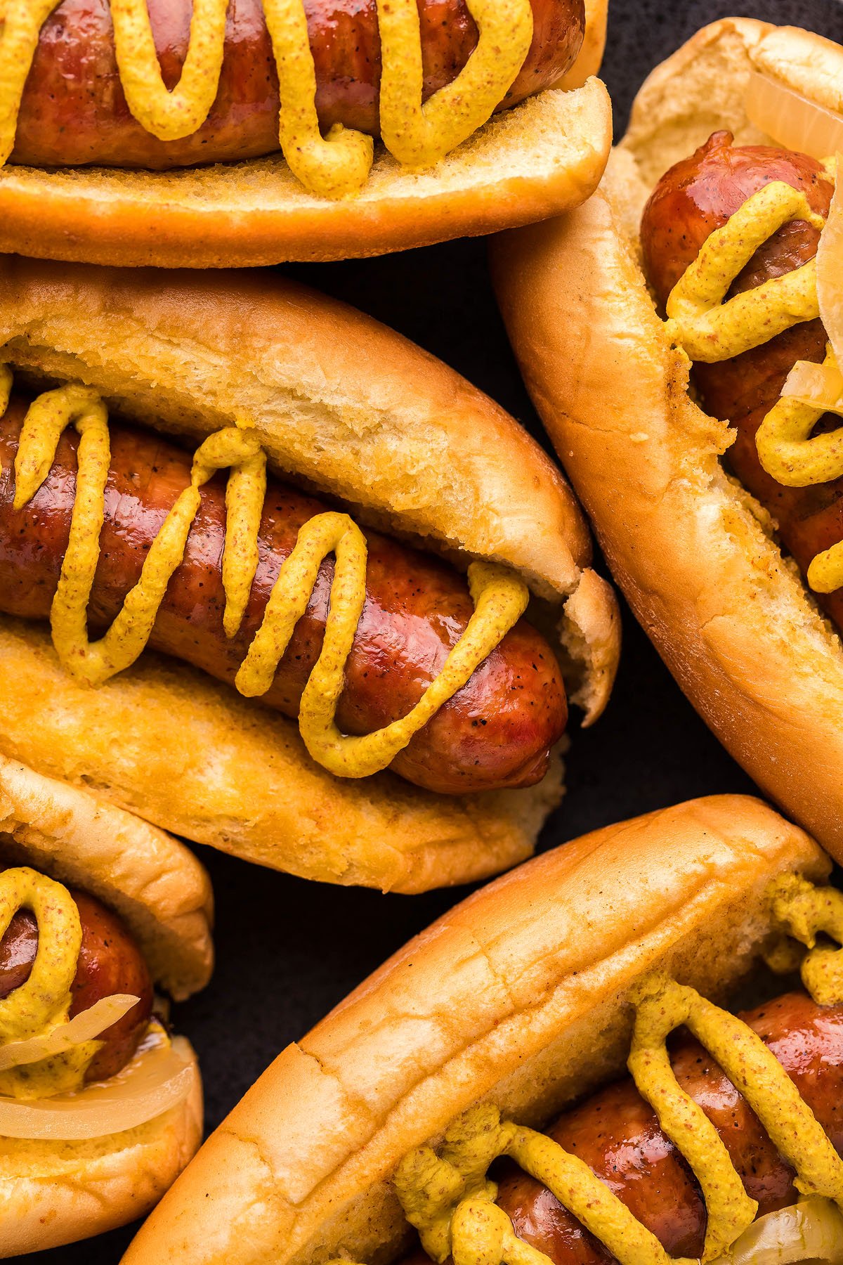 Several hotdogs in fluffy buns with thick drizzles of mustard.
