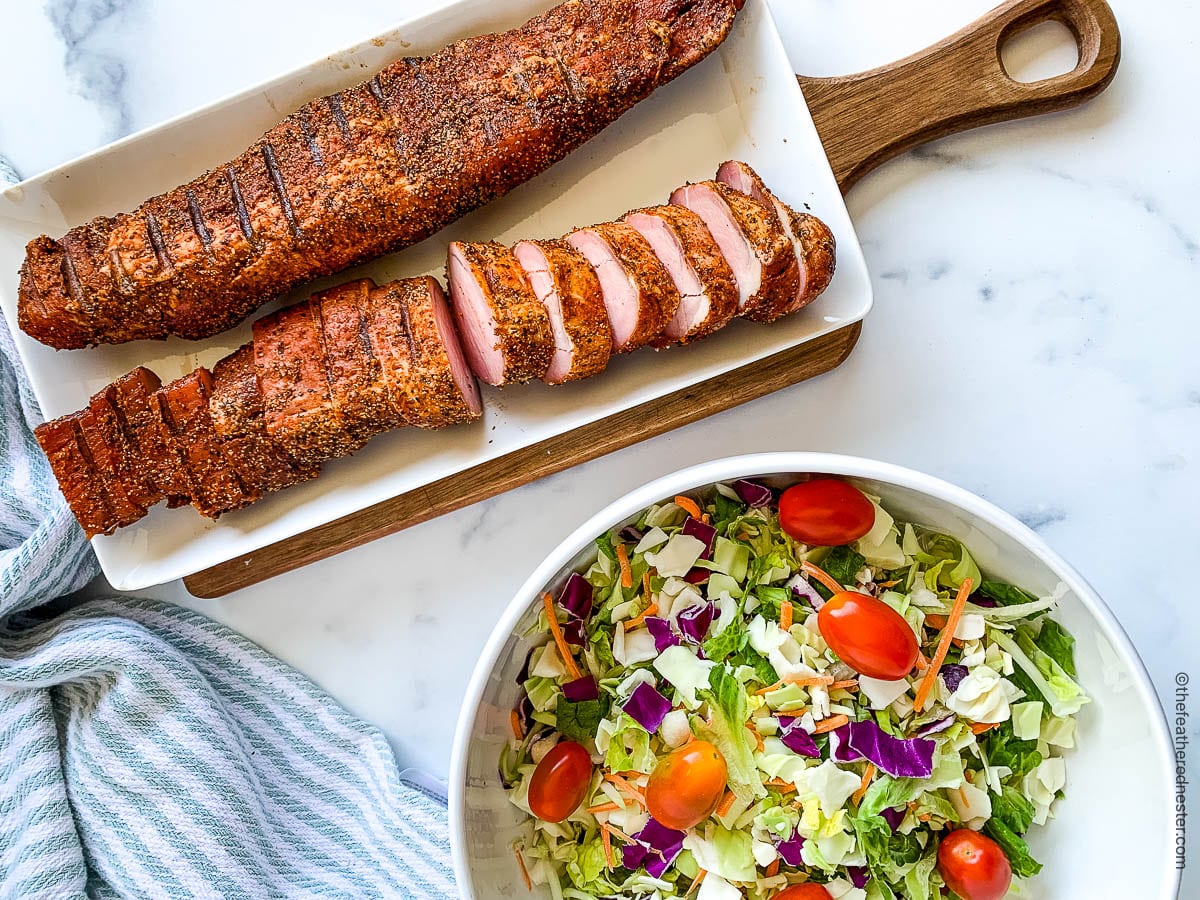 A whole and sliced smoked pork loin on a white serving platter next to a bowl of salad.