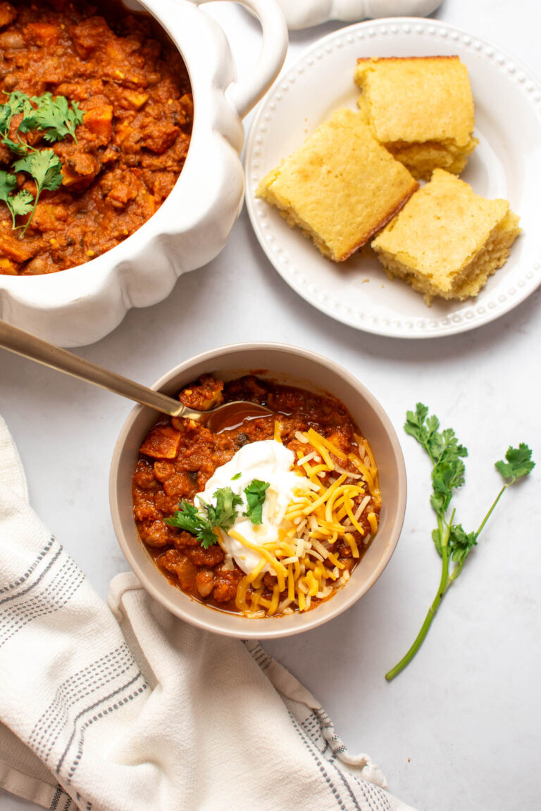 Bowl of vegetarian pumpkin chili in bowl with toppings next to a plate of cornbread and pumpkin serve ware full of chili.