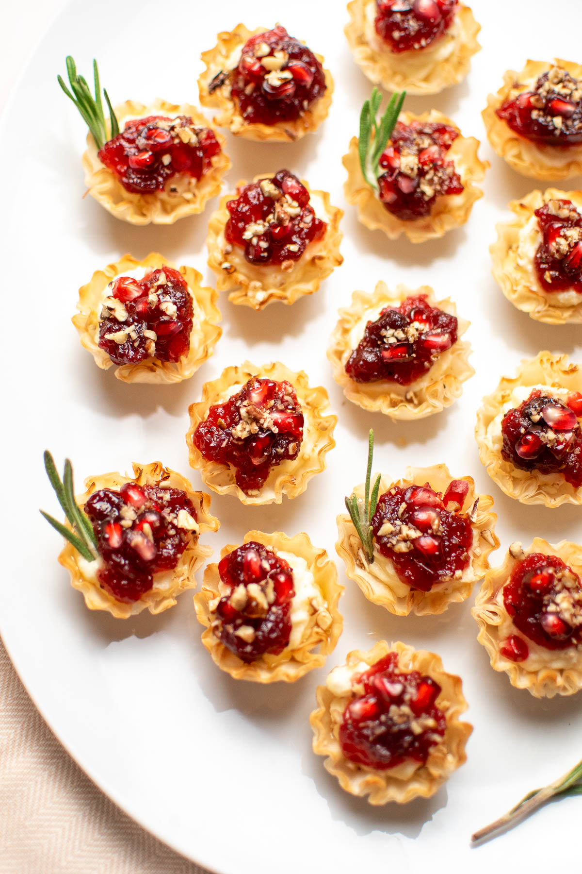 Cranberry brie bites with phyllo cups and rosemary on white platter.