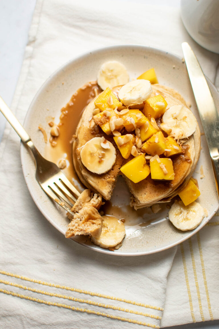 Stack of pancakes with mango and banana covered with brown syrup and fork with piece of pancake on the end.