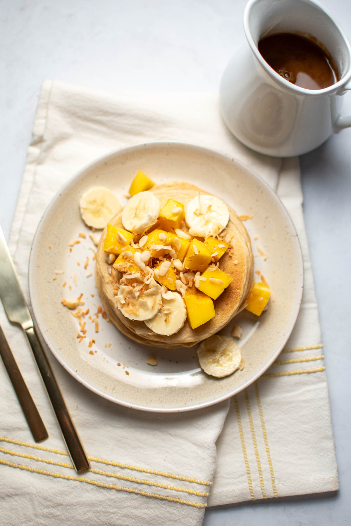 Plate of pancakes with slices of banana, mango, shredded coconut, and nuts on striped linen.