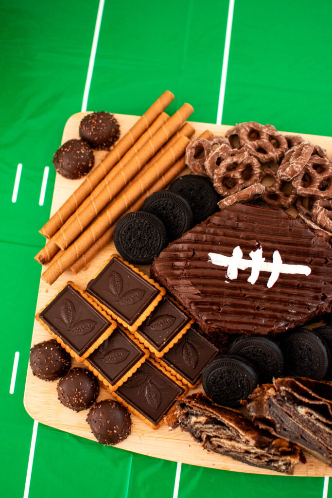 Wood cutting board with chocolate desserts on it and a football shaped cake in the middle.
