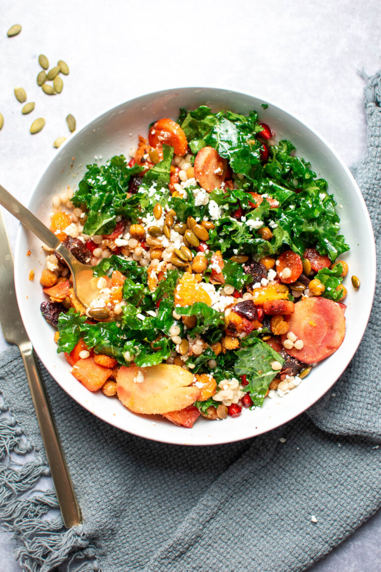 Gold fork in bowl with kale, roasted vegetables, pomegranate and pumpkin seeds on top of gray towel.