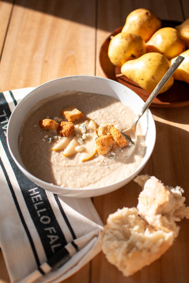Spoon in bowl of cauliflower and pear soup on table with pieces of bread and fresh pears nearby.
