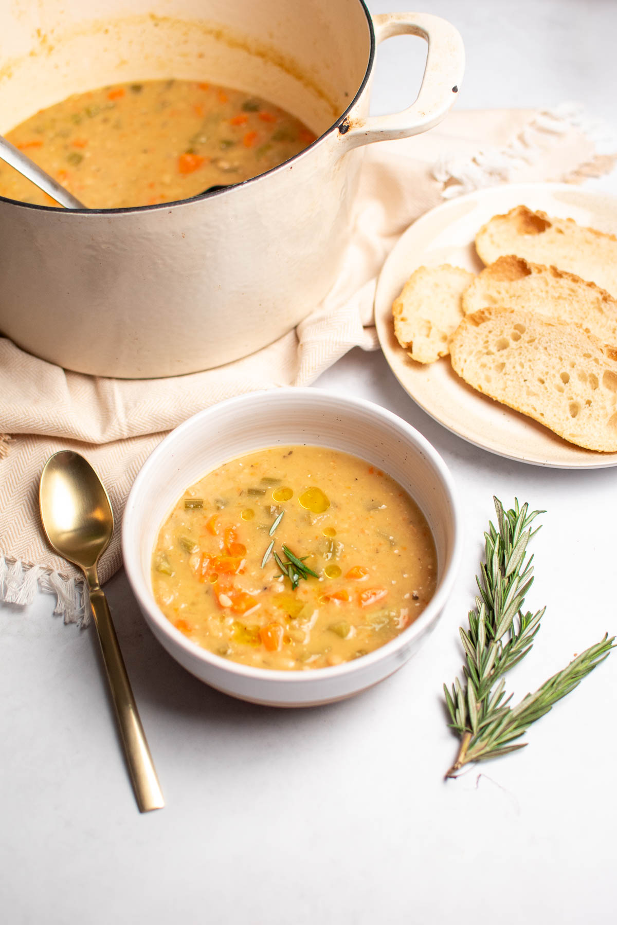 White bowl of white bean soup on table next to rosemary sprig, plate of bread, and soup pot.
