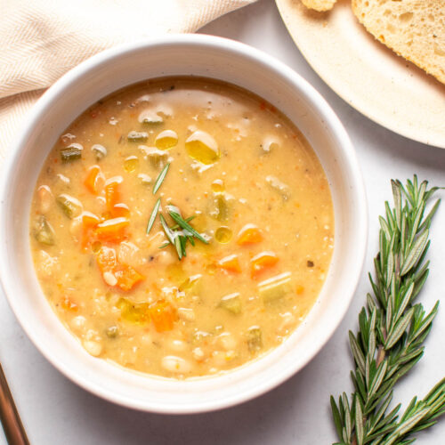 White bowl of soup with carrots and beans on table next to rosemary sprig.