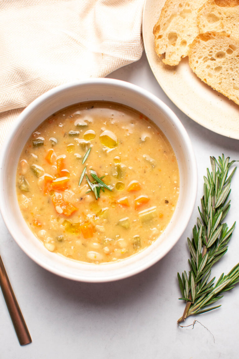 White bowl of soup with carrots and beans on table next to rosemary sprig.