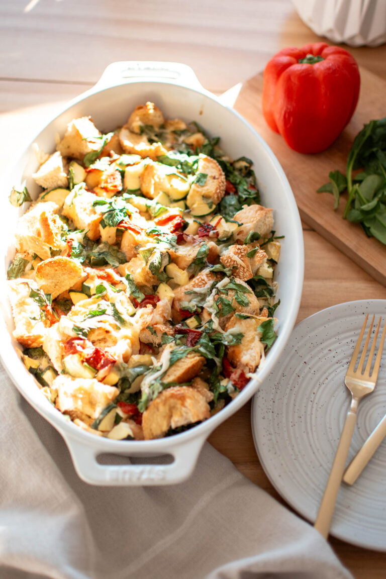 Veggie and egg strata in oval casserole dish with pepper and spinach nearby.
