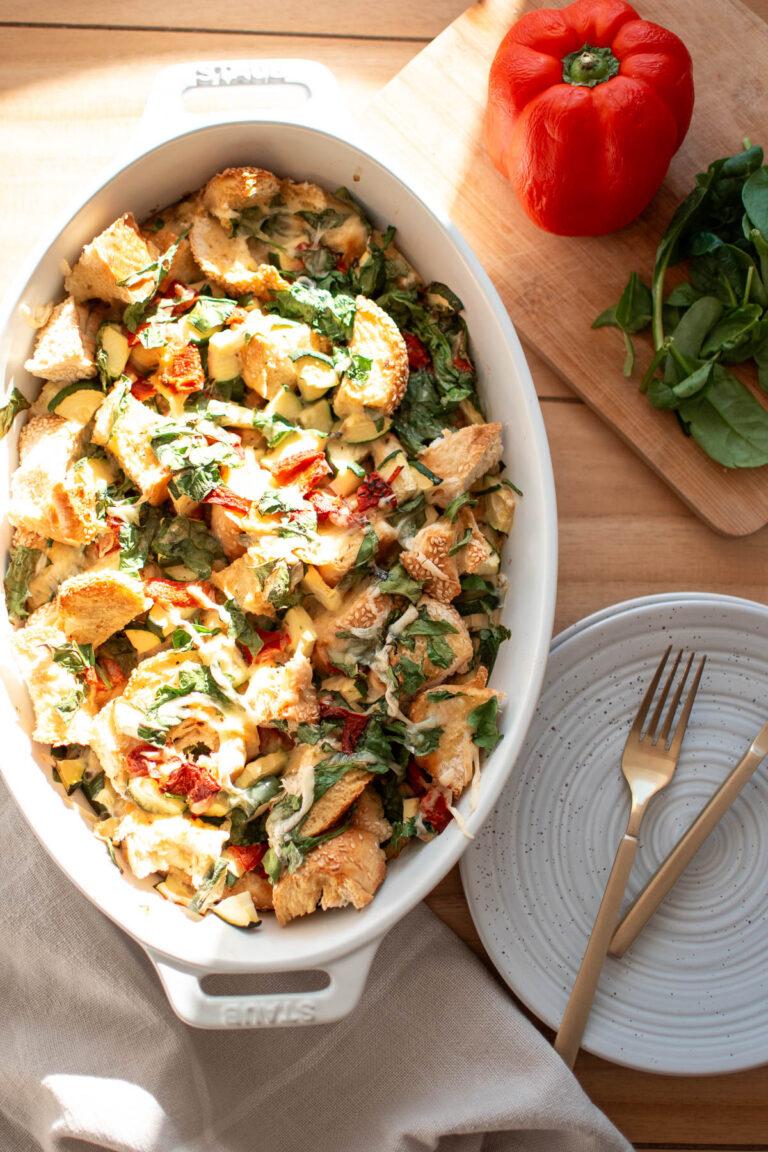 Veggie egg strata in oval dish on table with pepper, spinach, and plate.