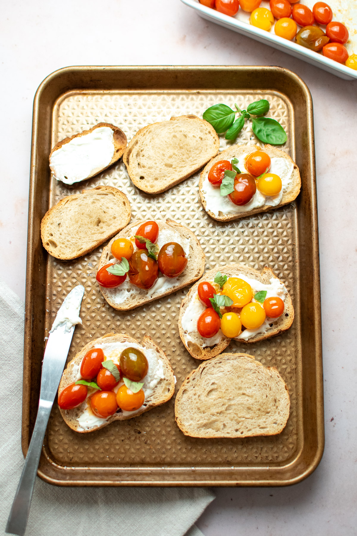 A sheet pan with sliced French bread being topped with whipped cream cheese, roasted tomatoes, and fresh basil leaves.