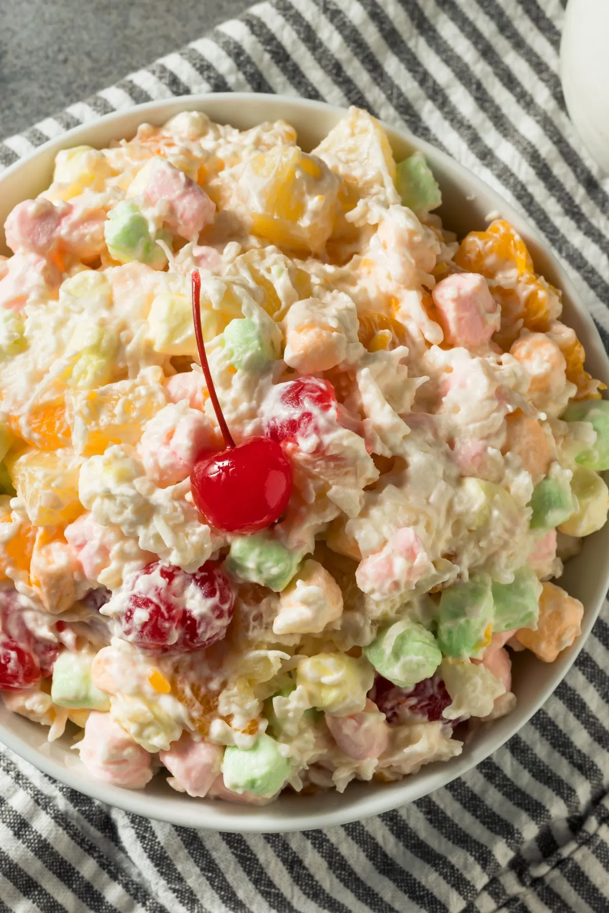Classic ambrosia salad with coconut and cool whip in a white bowl with a cherry on top.