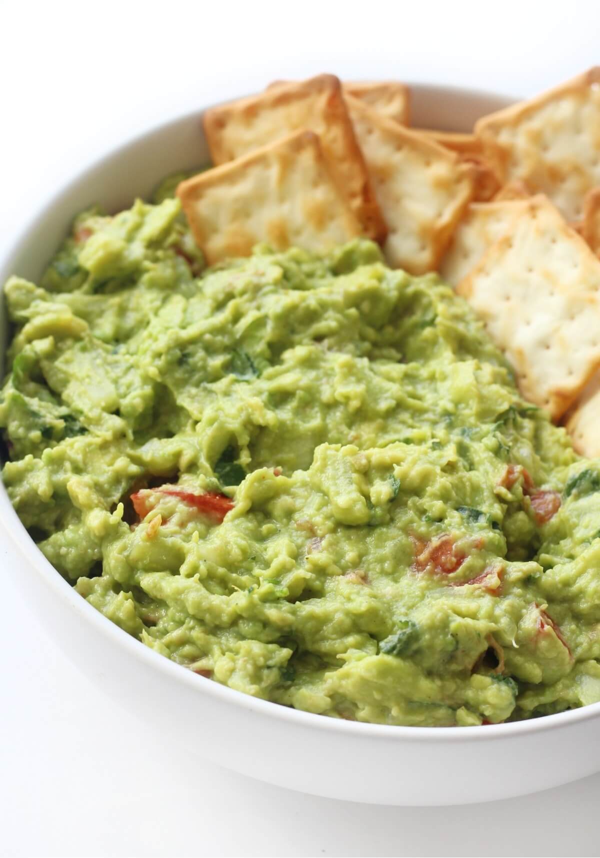 Homemade guacamole with tomatoes in a white bowl with crackers.
