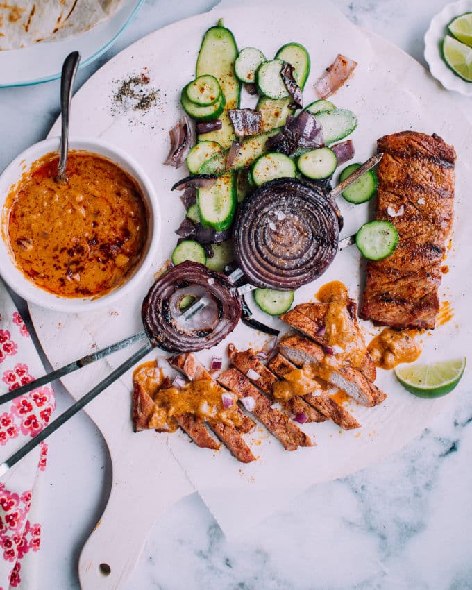Grilled pork tenderloin next to cucumber salad with a bowl of peanut sauce dressing.