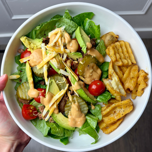 Woman holding white bowl with ingredients for high protein burger bowl.