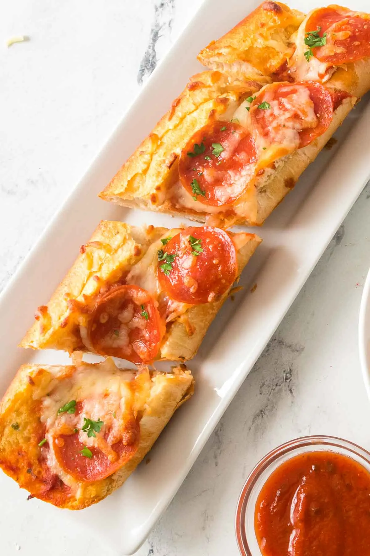 French bread pizza on a long white rectangular serving platter next to a small bowl of marinara dipping sauce.