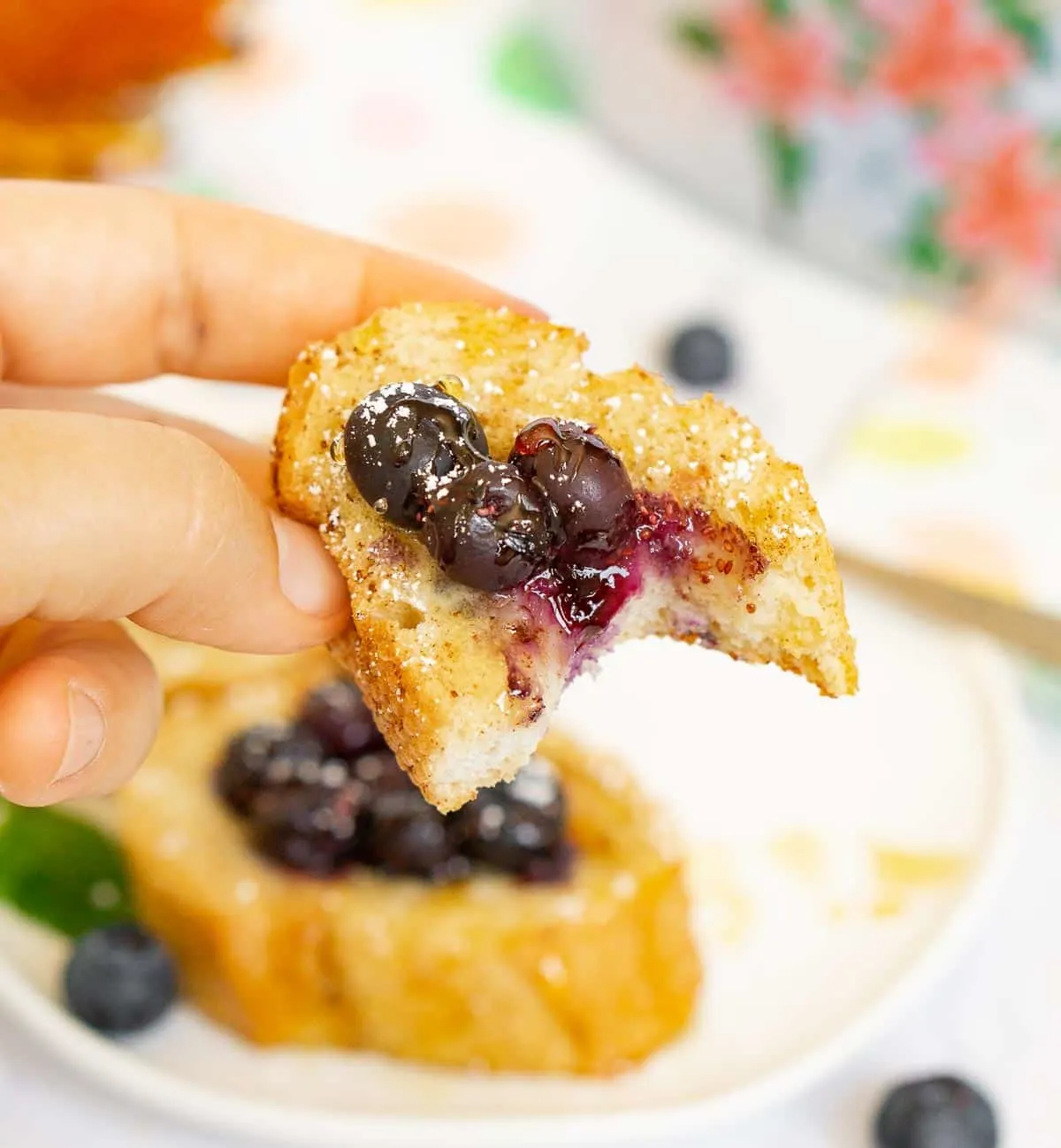 A woman lifts a French toast bite made with sliced French bread and blueberries from a white plate.