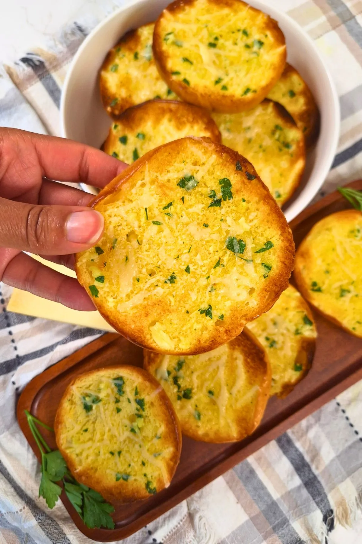 A woman holds a piece of hamburger bun garlic bread above a bowl and serving platter or more garlic bread.