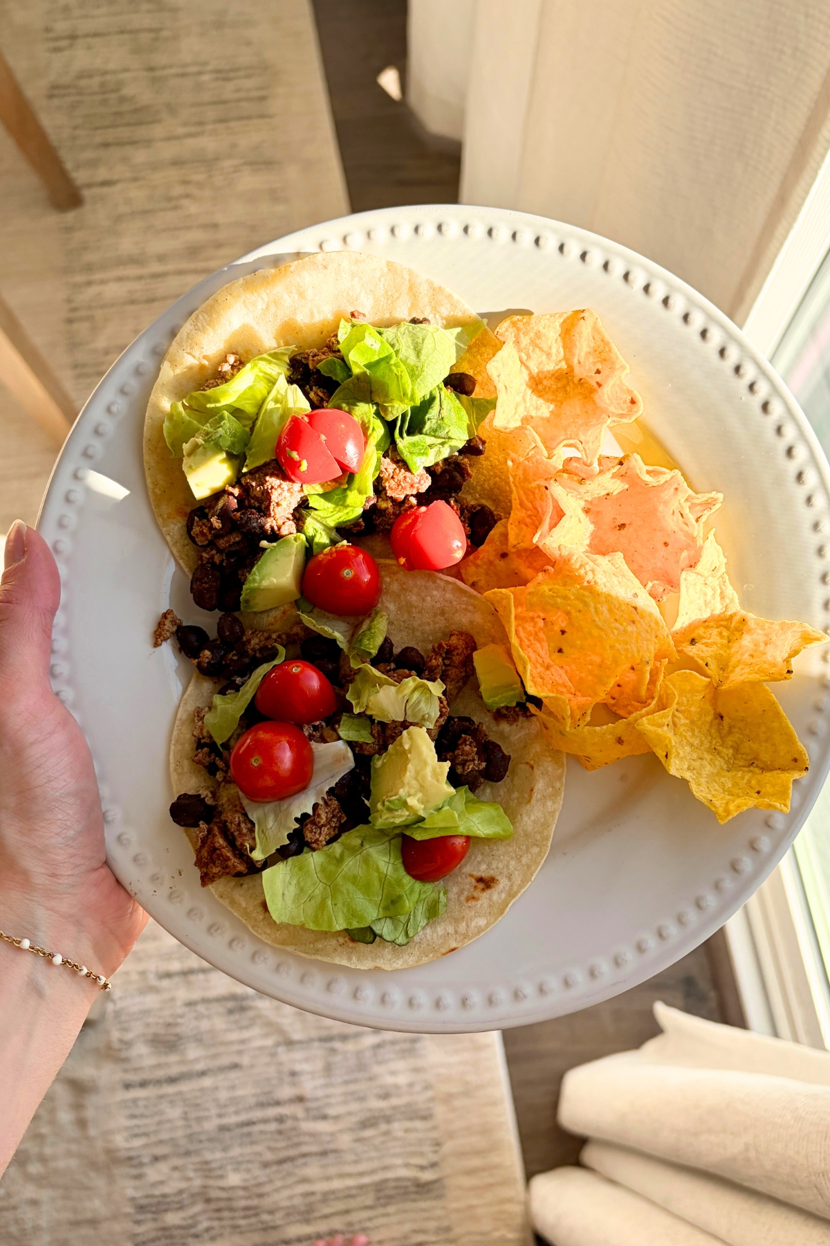 A woman holds a white plate that has two tofu black bean tacos with lettuce, tomato, and avocado, and some Tortilla scoop chips next to a dining room window.