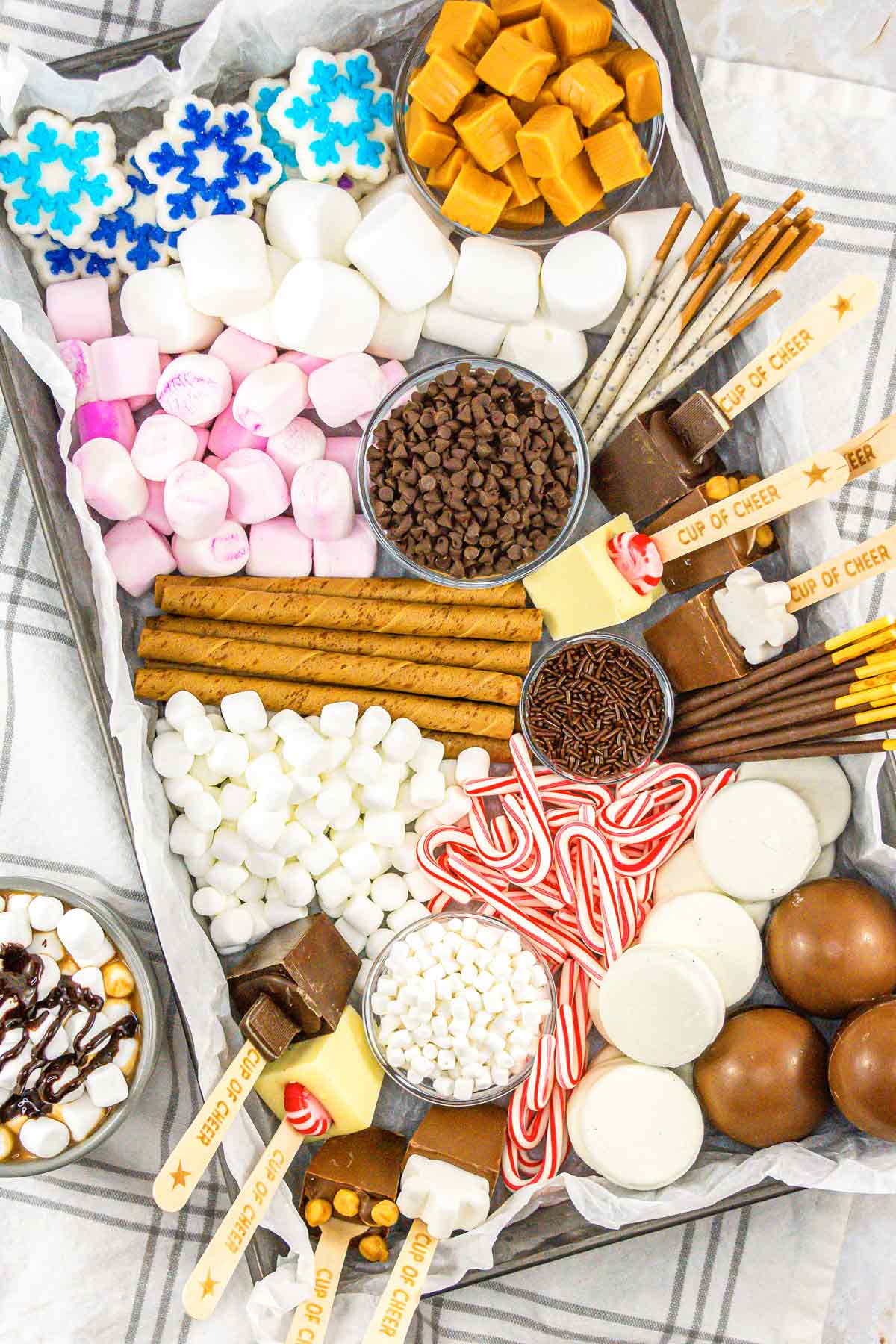 A hot chocolate charcuterie board with cookie sticks, marshmallows, peppermint, sticks, caramel, and chocolate.